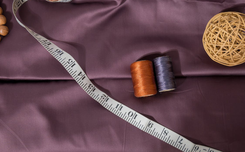 A measuring tape and two spools of thread rest on smooth mauve fabric, with a woven decorative ball in the corner.