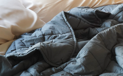 A rumpled charcoal weighted blanket rests across soft cream bedding, creating a warm, relaxed bedroom scene with gentle natural light.
